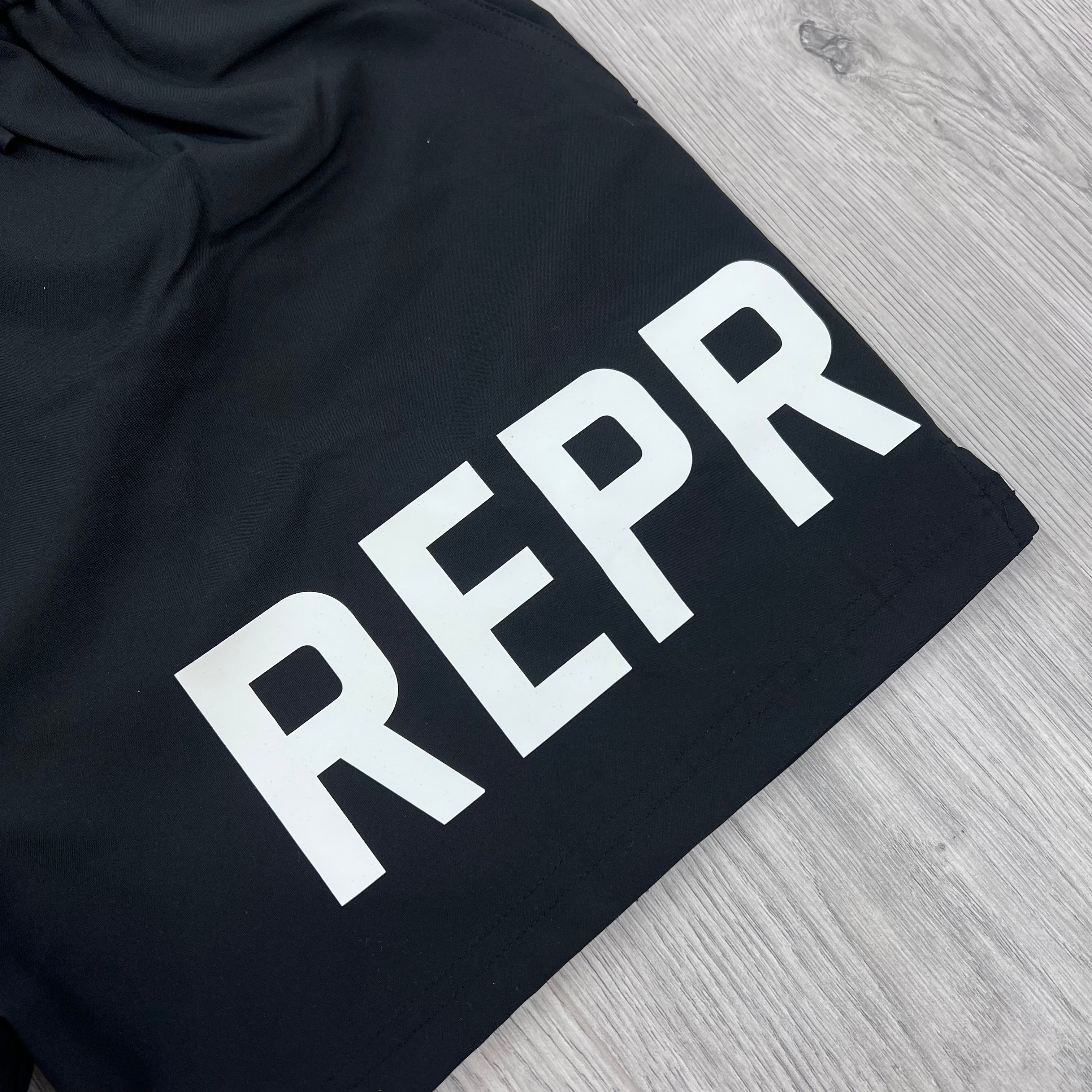 A close up view of the white logo on the front of the Represent Swim Shorts in Black.