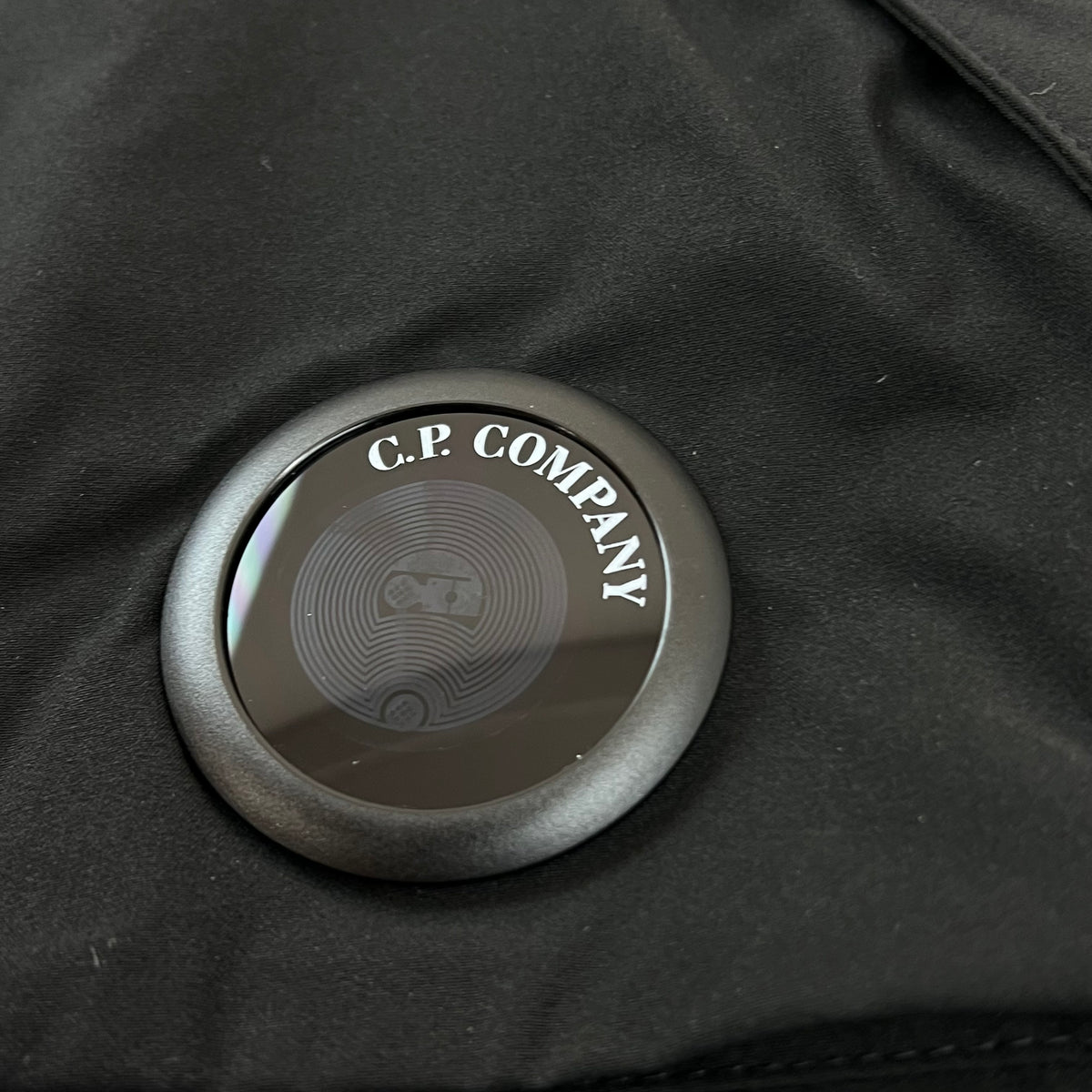 Close up view of the NFC-enabled lens logo detail on the CP Company Pro-Tek PrimaLoft Gilet in Black.