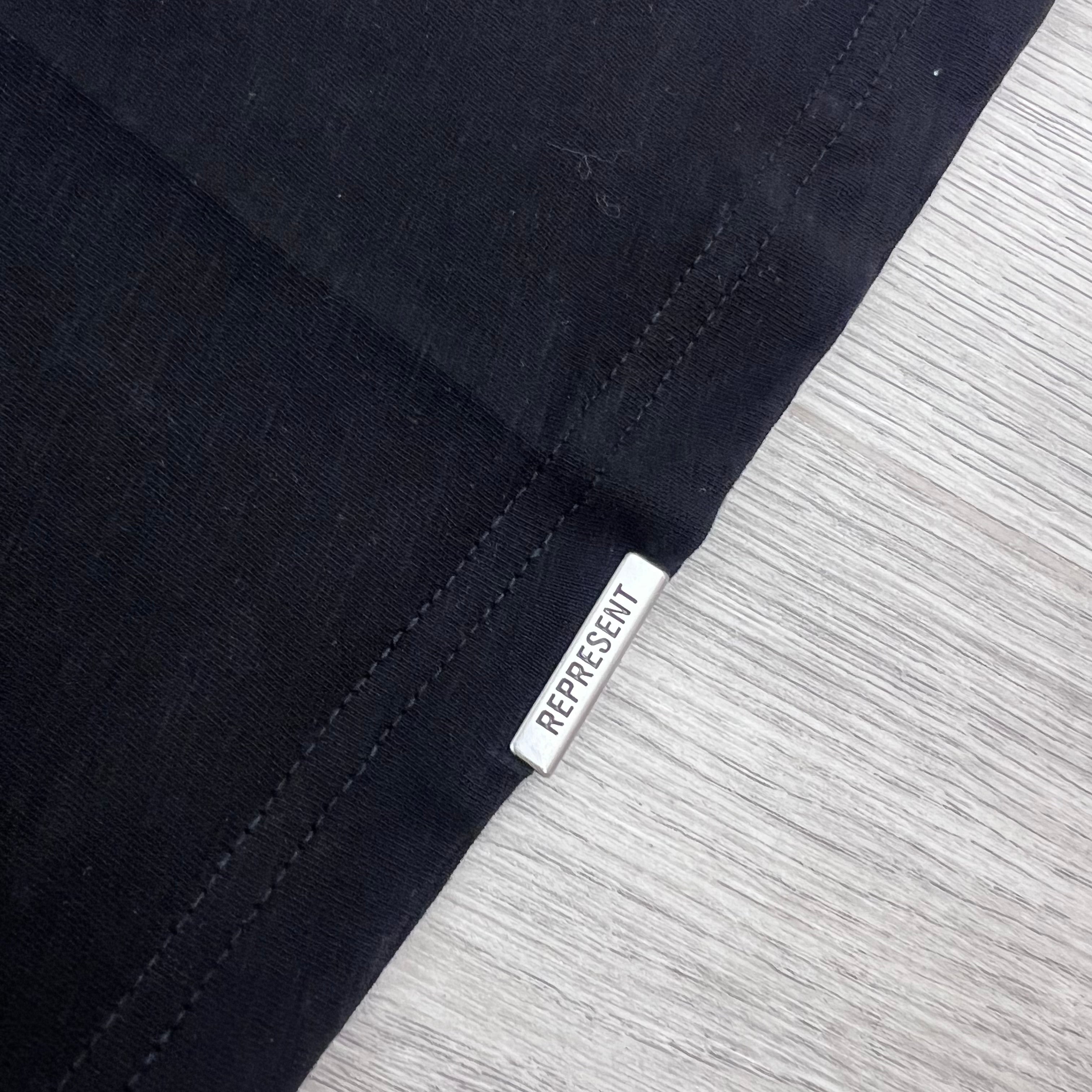 A close up view of the hardware detail on the Represent Owners Club T-Shirt in Jet Black.